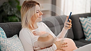 Mother and daughter sitting on sofa breastfeeding baby using smartphone at home
