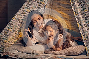 Mother and daughter are sitting in a teepee tent, reading stories with the flashlight. Happy family. photo