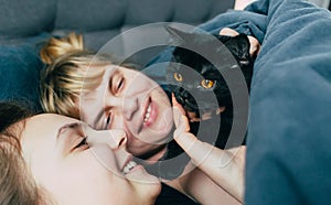 Mother and daughter sleeps together in a bed with a black cat. Family spending time together. Close up portrait of smiling woman h