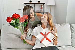 Mother and daughter smiling confident holding gift and flowers at home