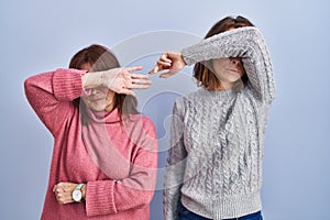 Mother and daughter standing over blue background covering eyes with arm, looking serious and sad