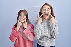 Mother and daughter standing over blue background smiling with open mouth, fingers pointing and forcing cheerful smile