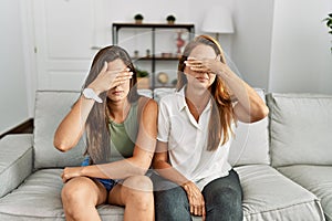 Mother and daughter together sitting on the sofa at home covering eyes with hand, looking serious and sad