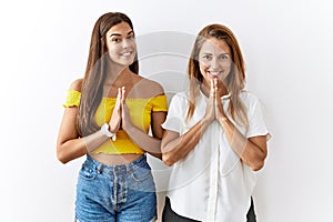 Mother and daughter together standing together over isolated background praying with hands together asking for forgiveness smiling