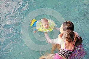 Mother and daughter training for infant baby boy in swimming suit floating in pool with safety by baby neck floats