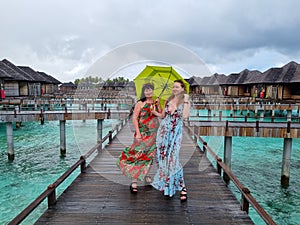 mother and daughter under umbrella, in matching tropical dresses standing together on wooden bridge pathway, women on vacations