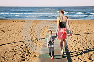 Mother and daughter walking by wooden flooring on sand beach at seaside. Summer family vacation. Children care and support by pare