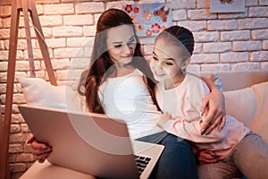 Mother and daughter watching movies on laptop under lampshade at night at home.