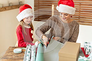 Mother and daughter wearing santa hats having fun wrapping christmas gifts together in living room. Candid family christmas time.