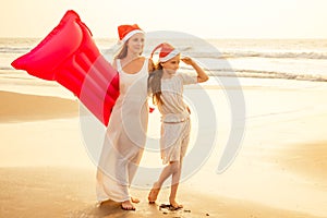 Mother and daughter in white dress and red Santa Claus hat having fun walking by summer sunset beach with air mattress