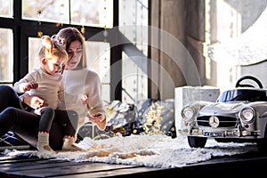 Mother and daughter in white sweaters sit on the floor near a large window and examine the New Year`s garland