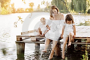 Mother day. Happy young mom with little girls sitting on wooden bridge near lake, pond on summer. Mother and two child