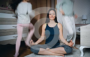 Mother doing yoga meditation exercise indoors at home, daughters playing.