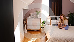 Mother dresses baby girl on bed against the window. Children's room, bedroom, mother takes the baby in her arms.