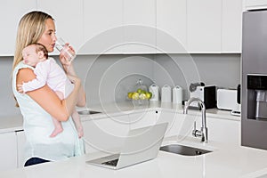 Mother drinking water while carrying baby in kitchen