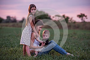 Mother embraces son and daughter, sitting on grassy field as twilight settles. Mothers day