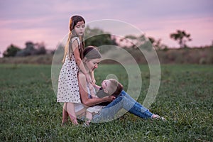 Mother embraces son and daughter, sitting on grassy field as twilight settles. Mothers day