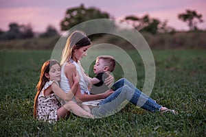 Mother embraces son and daughter, sitting on grassy field as twilight settles. Mothers day