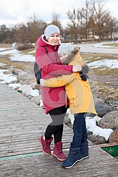 Mother embracing her frozen daughter, adult woman and young girl stand on quay at winter cold weather, people dressed warm clothes