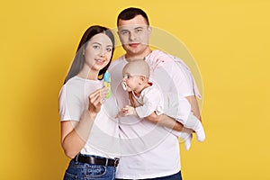 Mother, father and baby child posing isolated over yellow background, mommy with beanbag in hands, family wearing casual white t