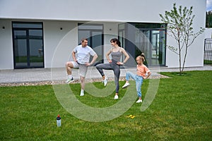 Mother, father and daughter with hands on hips doing leg exercise in yard