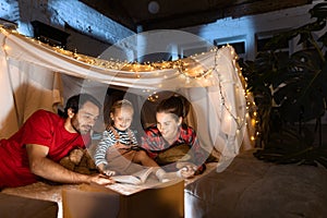 Mother, father and daughter lying inside self-made hut, tent in room in the evening and reading book. Family time