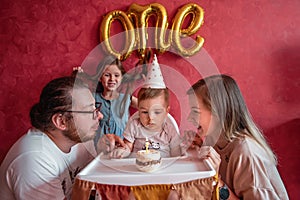 Mother father help blow out candle to one-year-old son on birthday cake. Happy family celebrating