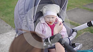 Mother feeding baby with orange spoon, child sitting in stroller outside the house