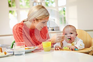 Mother Feeding Baby Sitting In High Chair At Mealtime