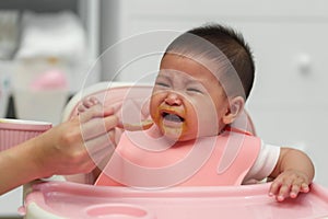 mother feeding food to her crying infant baby with spoon, refuse to eat