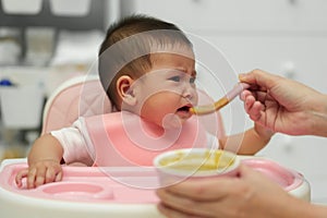 mother feeding food to her crying infant baby with spoon, refuse to eat