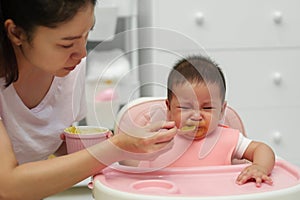 mother feeding food to her crying infant baby with spoon, refuse to eat