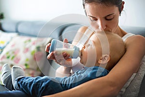 Mother, feeding her baby boy from bottle, sitting on the couch a