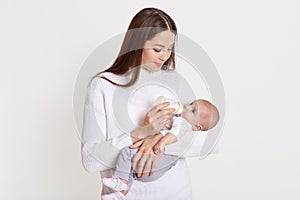 Mother feeding her baby from feeding bottle, dark haired female wearing sweater hold baby in hands and looking at her kid, posing