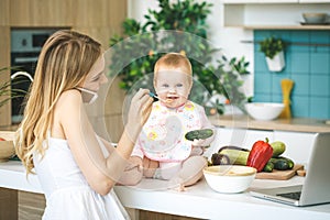 Mother Feeding Her Baby Girl with a Spoon. Mother Giving Food to her Child at Home. Baby food. Working home. Using phone