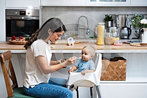 Mother feeding her baby son or daughter with spoon. Mother giving healthy food to her adorable child at home. Lovely mommy and her