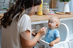 Mother feeding her baby son or daughter with spoon. Mother giving healthy food to her adorable child at home. Lovely mommy and her