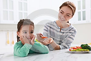 Mother feeding her daughter in kitchen. Little girl refusing to eat vegetables
