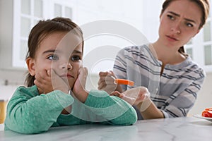 Mother feeding her daughter in kitchen. Little girl refusing to eat vegetables