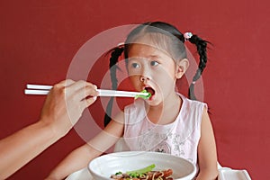 Mother feeding noodle for her daughter on high chair against red wall background