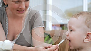 Mother feeds her young son with ice cream from a spoon. They are sitting in a street cafe. The boy really likes the