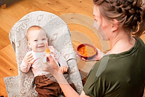 Mother feeds laughing baby with vegetable puree from plastic spoon at home