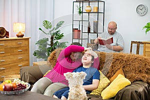 Mother is fooling around with her son on the sofa and father is reading a book.