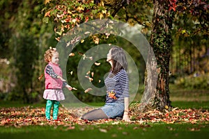 Mother and girl having fun under tree with autumn leaves in the park, blond cute curly girl, happy and young family