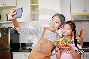 Mother and girl preparing salad and taking a selfie by smartphone for social media. Leisure activity