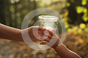 Mother giving her daughter glass of fresh water in forest on sunny day, closeup