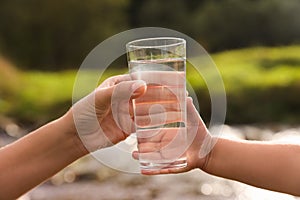 Mother giving her daughter glass of fresh water near stream on sunny day, closeup