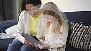 Mother or grandmother is sitting on sofa and showing to little girl digital technology