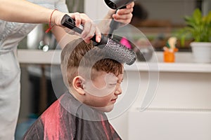 mother with a hairdryer in her hand in a light blue dress is doing her son`s hair at home during the second period of quarantine