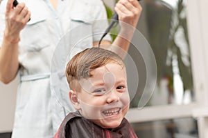 mother with a hairdryer in her hand in a light blue dress is doing her son`s hair at home during the second period of quarantine